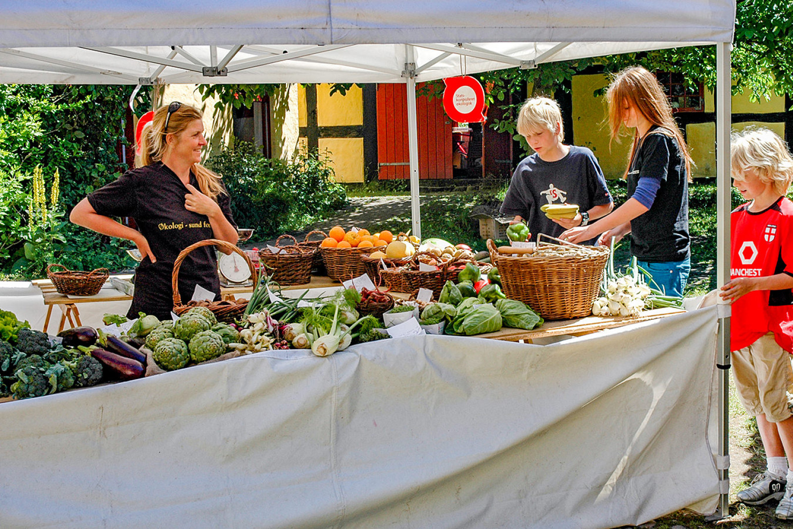 Tre personer står ved en madbod og ser på frugt og grønt. Sælgeren står smilende og ser på.