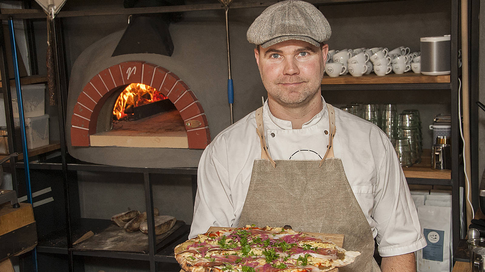 Ole Olesen står med en pizza foran en tændt stenovn.
