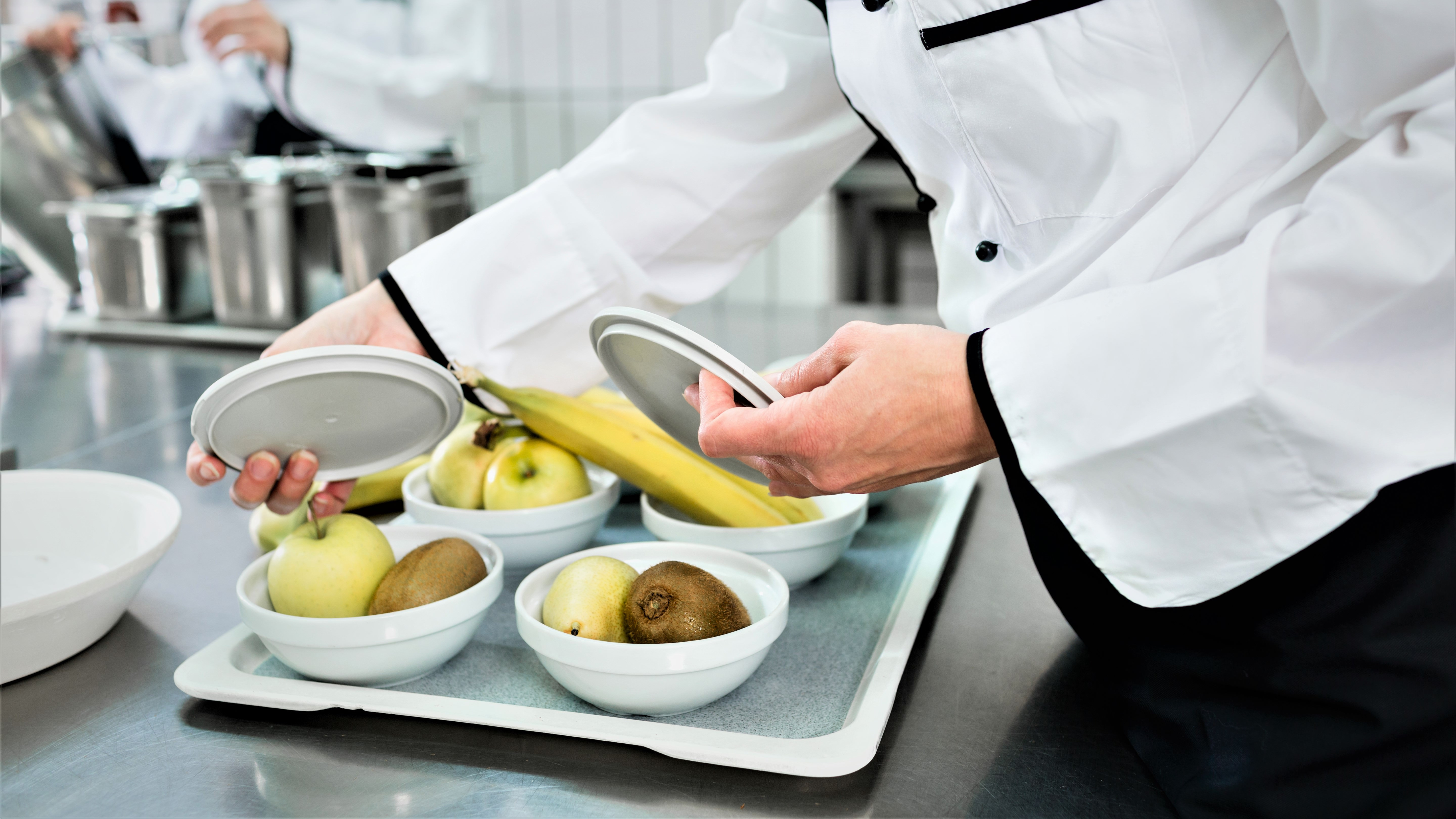 En kok er ved at anrette skåle med frugt; bananer, kiwier og æbler, i et køkken.