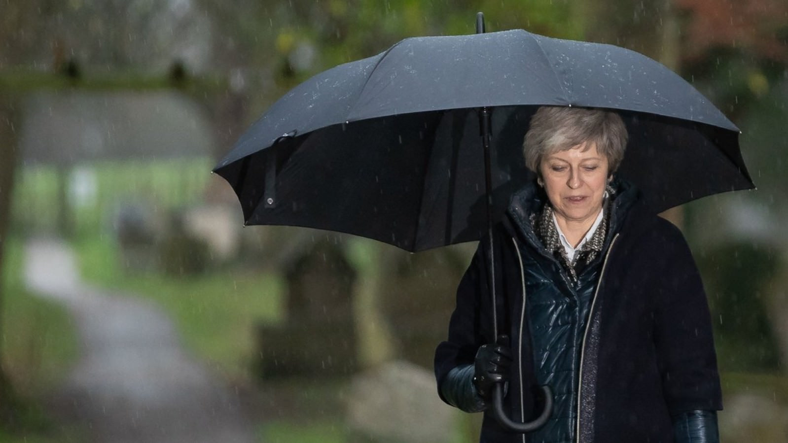 Theresa May går iklædt sort i regnvejr og holder en paraply, mens hun kigger ned i jorden