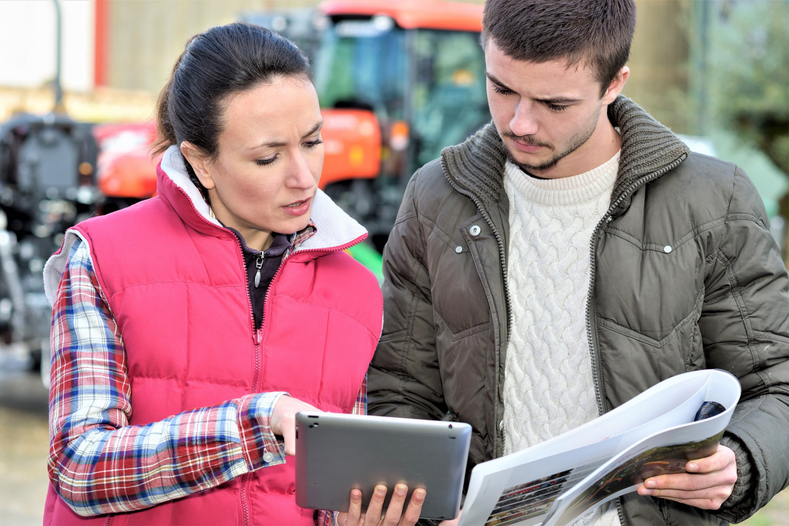 To personer står og kigger ned i et magasin og en tablet med traktorer bag sig.