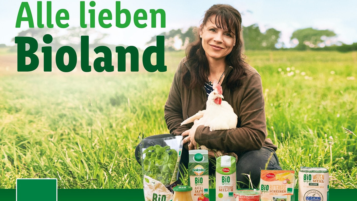 Et kampagnebillede for Bioland, der viser en kvinde sidde på marken med en høne - foran hende er en række Bioland-produkter