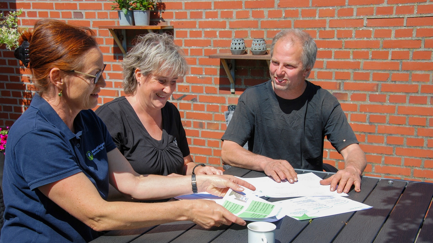 Solvejg Horst sidder ved et havebord og viser Tina og Jan Bolding nogle papirer
